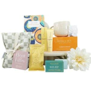 Assorted self-care gift set including tea, heat pillow, lotions, bath products, a checkered tote, and a white flower.