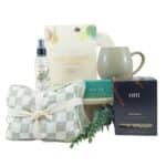 Self-care gift set including a checkered pillow, spray bottle, tea, green box labeled RELIEF, a dark mug, and a leafy sprig.