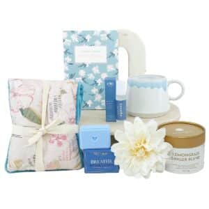 Spa gift set with blue floral box, ceramic mug, candle, soap, and wrapped floral towel arranged with a white flower.