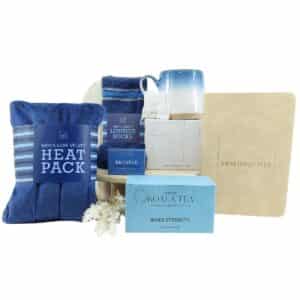 Blue-themed gift set including a heat pack, velvet lounge socks, mug, tea (Koala Tea Inner Strength), shower steamer, puzzle book, and dried flowers.