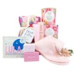 Pamper gift set in pink floral and pastel packaging, including a heat pillow, eye mask, soaps, shower steamer, and a soft pink scarf/band.