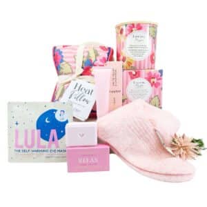 Pamper gift set in pink floral and pastel packaging, including a heat pillow, eye mask, soaps, shower steamer, and a soft pink scarf/band.