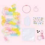 Flat lay of pastel kid craft items on a pink background: rainbow fringe garland, pink cord bracelet, pink beaded bracelet, cupcake-shaped cutout, sticker sheet with cute animals and treats, white resealable bag, small black beads, and a pink star-shaped clip toy with a matching lanyard.
