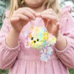Child in a pink dress holds a pastel fluffy pom-pom keychain with beads and kawaii charms outer scene visible in background.
