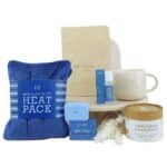Blue heat pack and spa gift set with towel, mug, candle, and bath products arranged on a round stone stand for a relaxing gift box feel