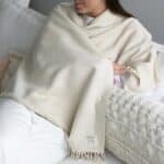 Woman sitting on a white bed, wrapped in a cream fringed blanket, with a tag reading 'HANDMADE with Love'.