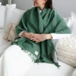 Person seated on a white sofa wearing a green fringed shawl over white clothing, hands clasped in lap.