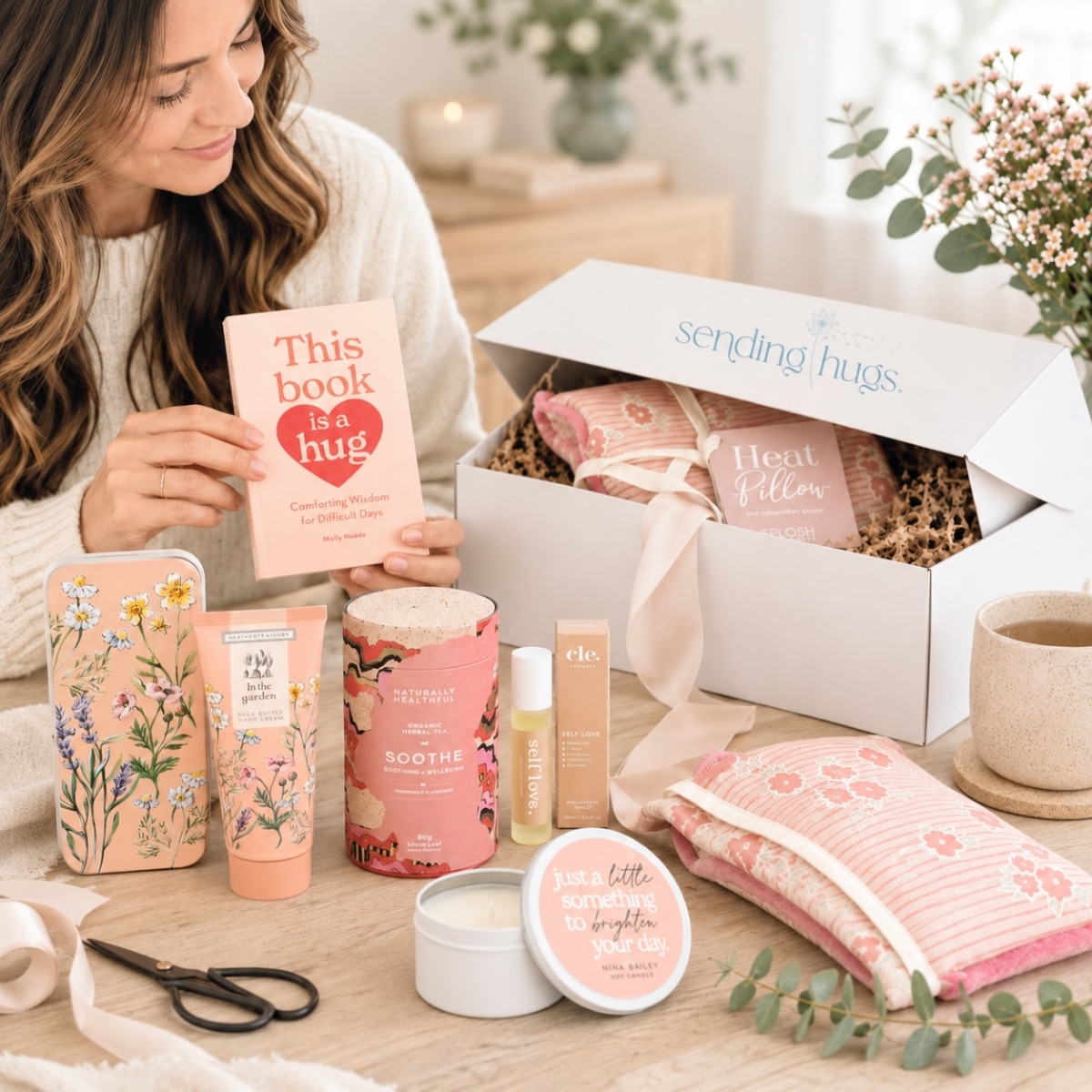 Woman unboxing a pastel pink self-care gift set on a wooden table, holding a book that reads 'This book is a hug.'
