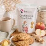 Bag of The Milk Pantry Cookie Mix in Ginger & Apple flavor on a kitchen table beside cookies, ginger, apples, and a mug of tea; cozy baking scene