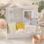 Gift box with a framed photo, beige decorative book, and small gift sachets on shredded filler, tied with a pale ribbon outside the box in a soft setting.