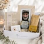 A gift box labeled "Sending Hugs" opened to reveal a photo frame, a beige book, chocolate bars, and small cards arranged with tissue paper and dried greenery.