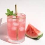 Pink watermelon cocktail in a ribbed glass with ice and a gold straw, garnished with mint leaves.
