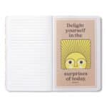 Open notebook with a peach card on the right page reading 'Delight yourself in the surprises of today' and a yellow sun illustration.