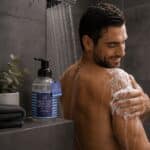Man in a shower applying lather to his shoulder; a labeled Mr Styln hand and body wash bottle sits on the shelf nearby with a plant and folded towels beside it (informative).