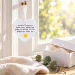 Sending Sunshine acrylic heart ornament hanging beside a bright window with soft linen and a Sending Hugs gift box.